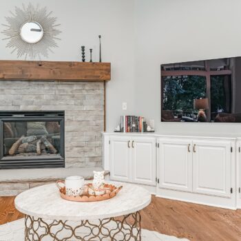 A living room with a tall limestone fire place, round marble coffee table, and white cabinet entertainment center.