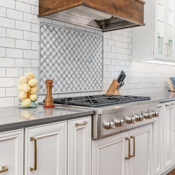 Kitchen remodel using white cabinetry with gold handles, white wall tile, and wood stove ventilation designed by Farha Home Trends.